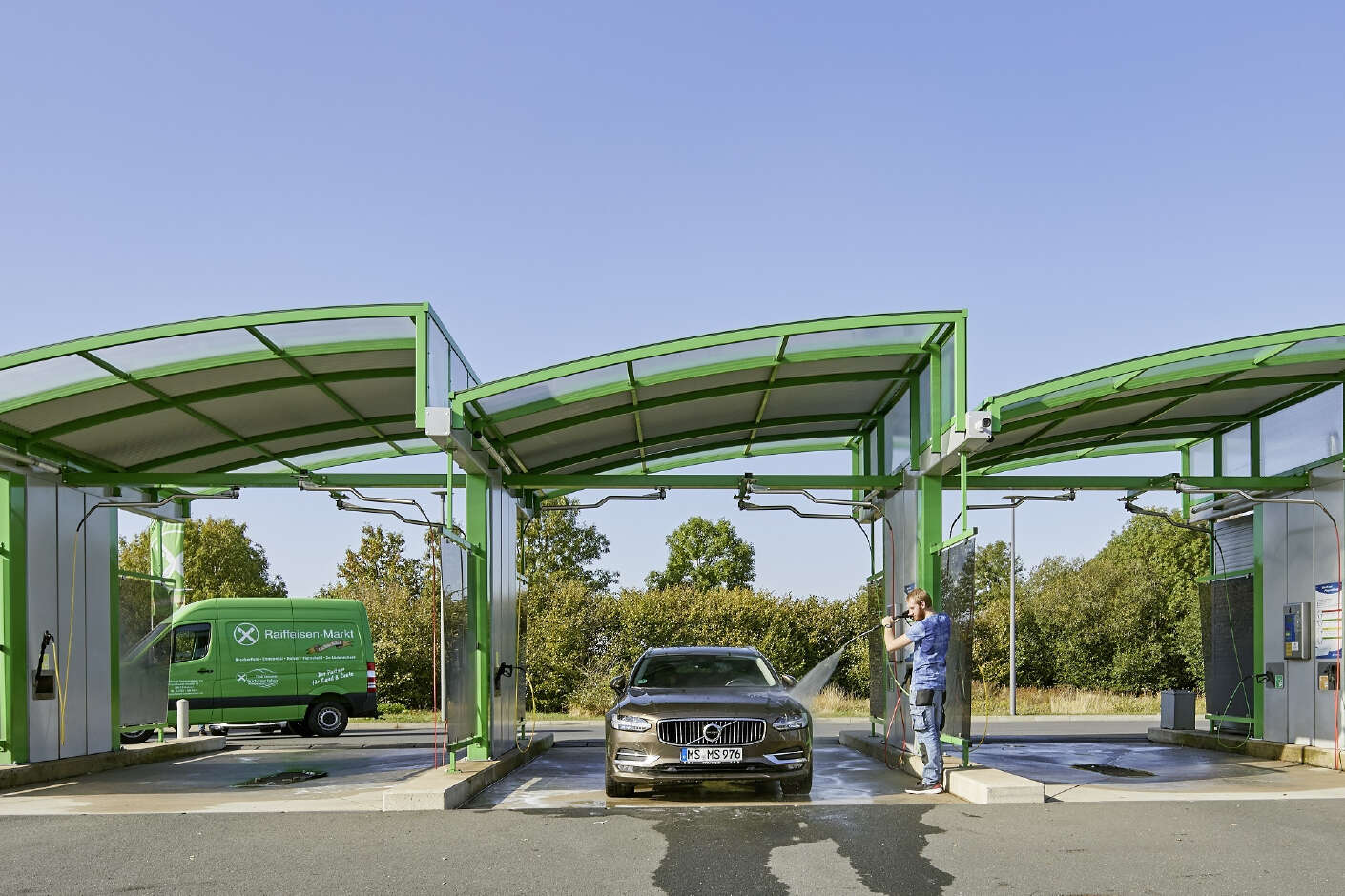 Drei Waschboxen nebeneinander. Das Stahltragwerk der Waschboxen ist grün. In der mittleren Waschbox wird ein Auto mit einem Hochdruckreiniger gewaschen.
