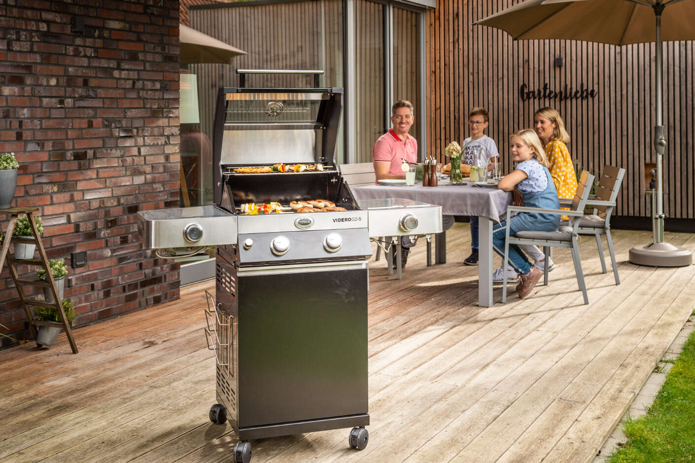 Grill mit Grillgut im Vordergrund eine Familie im Hintergrund auf einer Terrasse