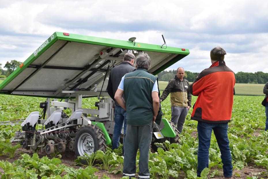 Der Feldroboter Farmdroid FD20 steht auf einem Acker, ringsherum stehen mehrere Personen. 