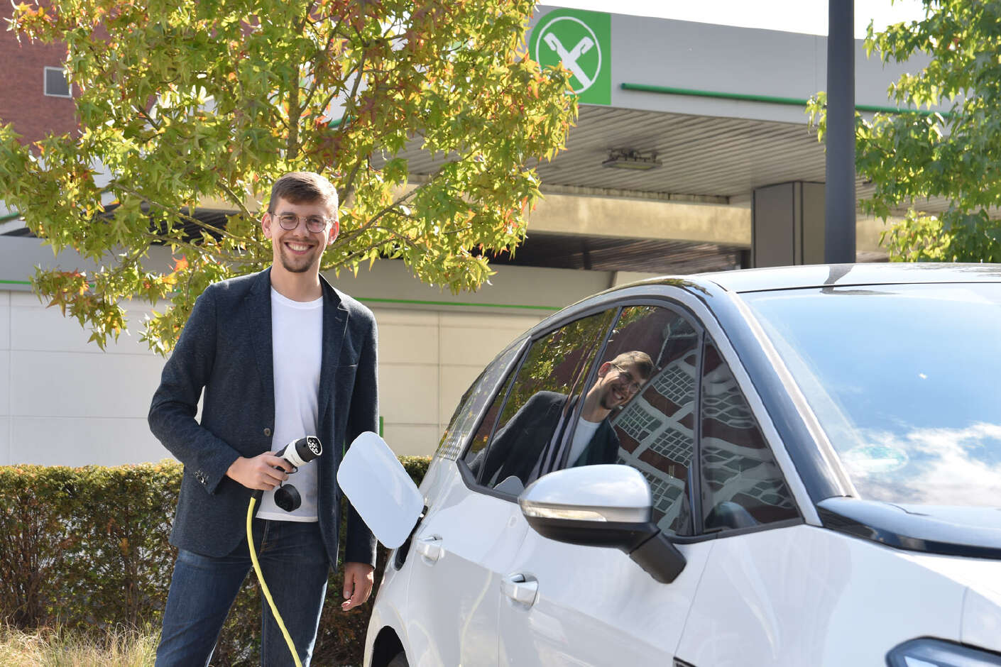 Ein Mann steht lächelnd neben dem weißen E-Auto, den Tankschlauch in der Hand. 