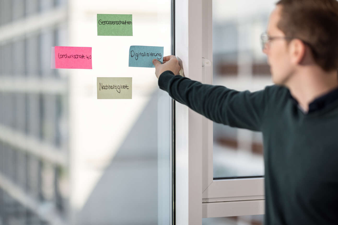 Mitarbeiter klebt Post-Its mit dem Begriffen Genossenschaften, Digitalisierung, Nachhaltigkeit %26 Landwirtschaft 4.0 auf eine Scheibe