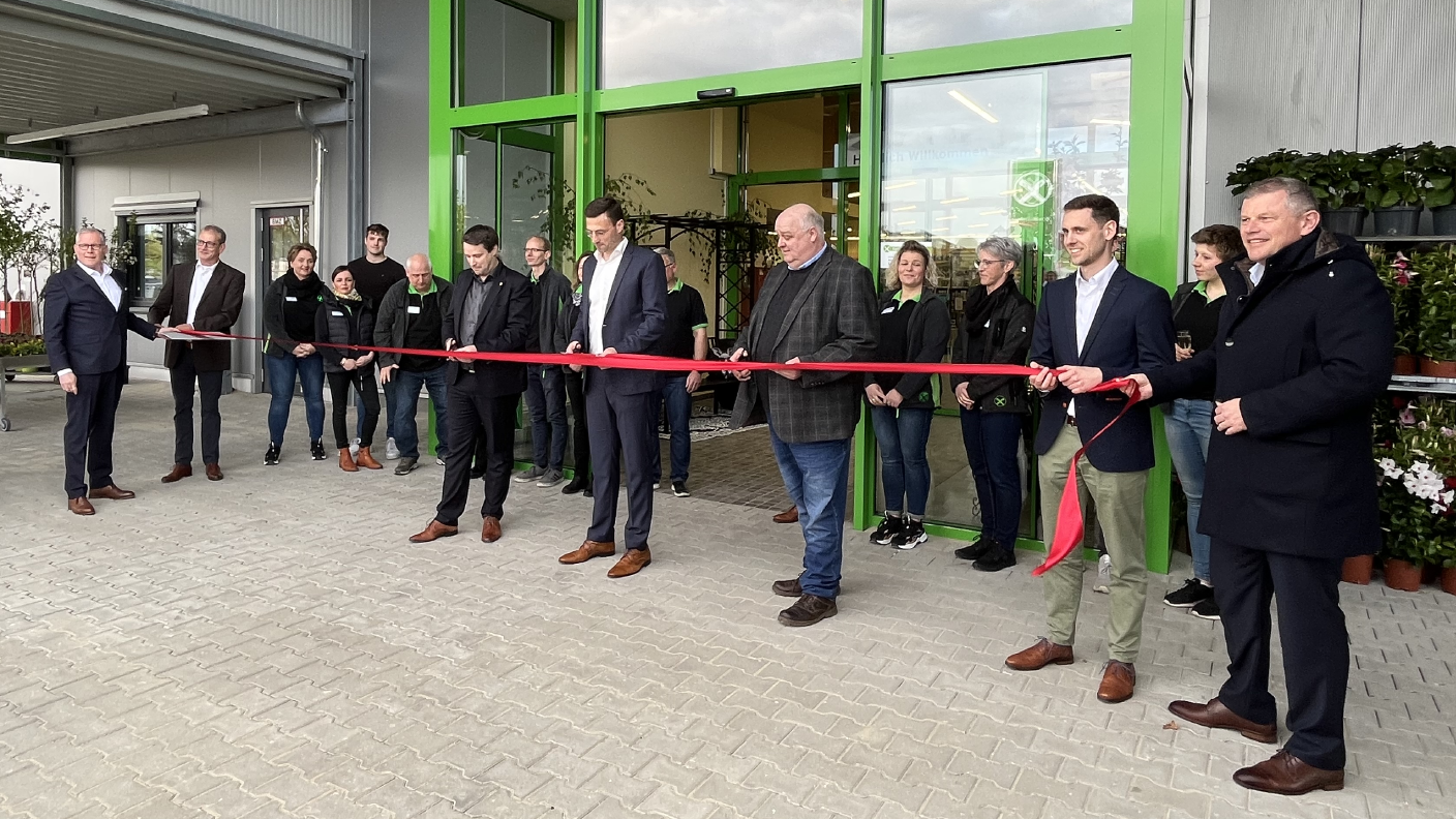 Mehrere Menschen vor dem Eingang des Raiffeisenmarktes in Uetze. Die Menschen halten gemeinsam ein großes rotes Band in den Händen. Der Mann in der Mitte der Menschen hält eine Schere in der Hand und setzt zum Durchschneiden des roten Bandes an.