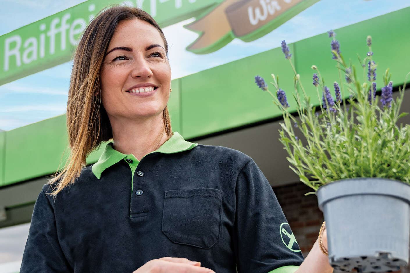 Eine lächelnde Mitarbeiterin steht vor einem Raiffeisenmarkt mit Blumen in der Hand. 