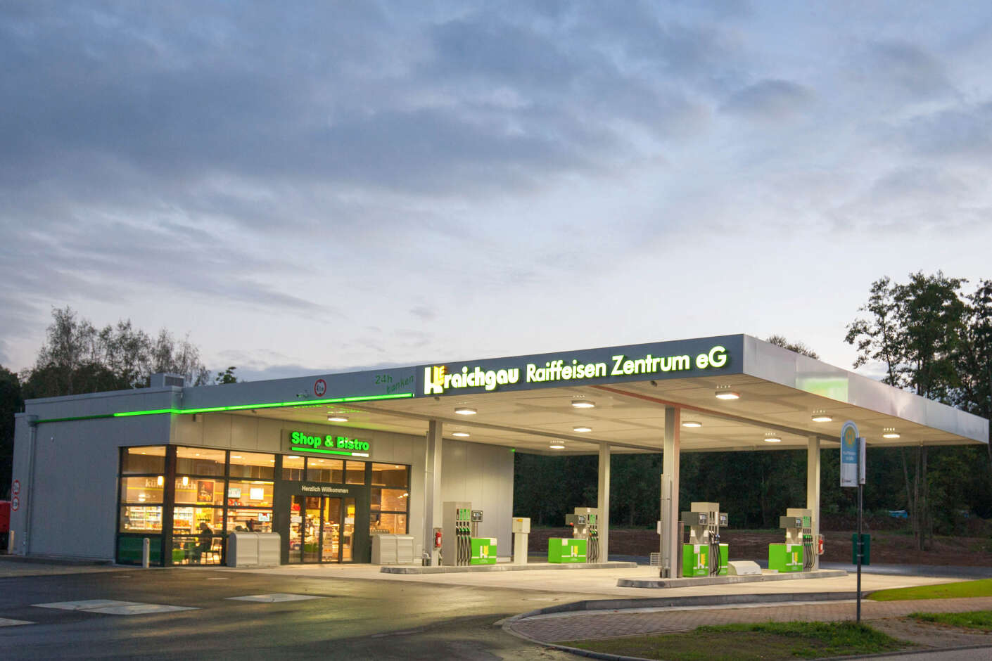 Beleuchtete Tankstelle in der Dämmerung mit acht Tankstellenfeldern, Shop und Bistro. Am Dach und Eingang befindet sich grün leuchtende Schrift.