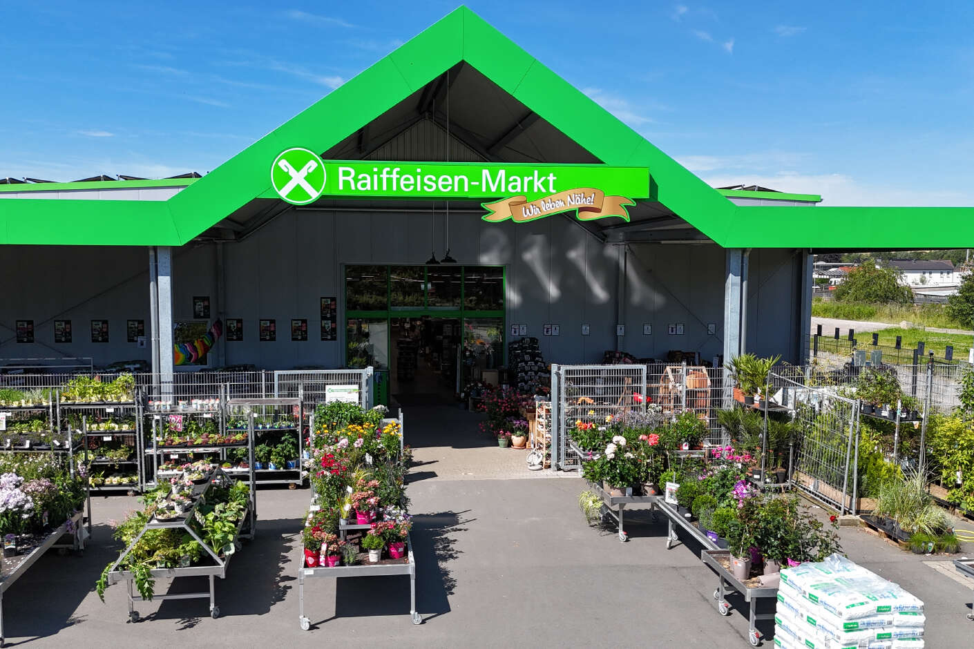 Pflanzenabteilung des Raiffeisenmarktes. Die Pflanzenabteilung befindet sich unter einem grünen Giebeldach mit Raiffeisenmarkt-Werbung. 