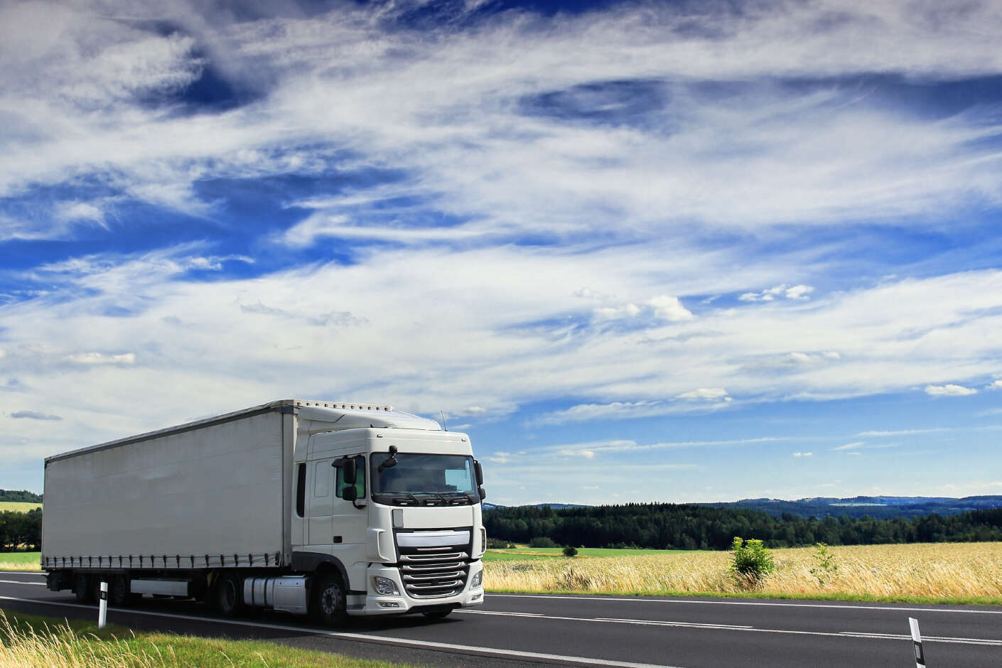 Ein weißer Lkw fährt durch grün, braune Felder. Der Himmel ist blau mit weißen Wolken.