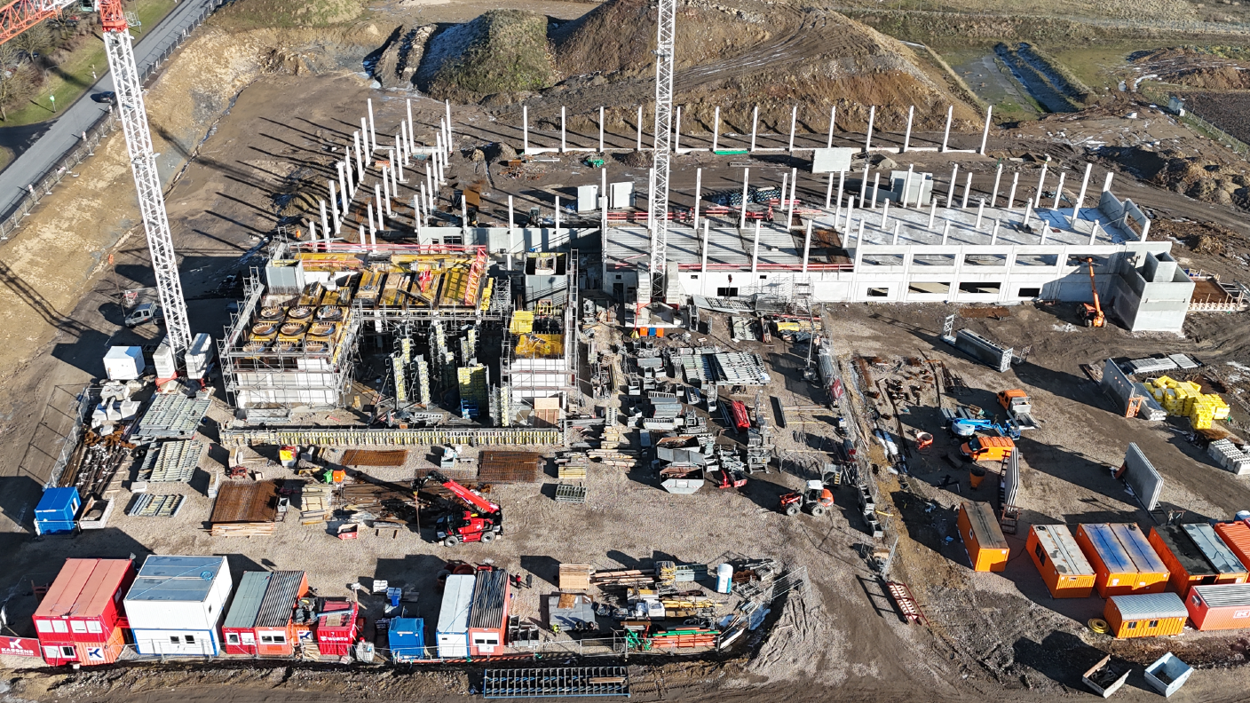 Baustelle mit Kran, Baucontainern und etlichen Baustellenfahrzeugen von oben mit einer Drohne fotografiert. Die ersten Betonstützen und massiven Wände sind fertig betoniert. 