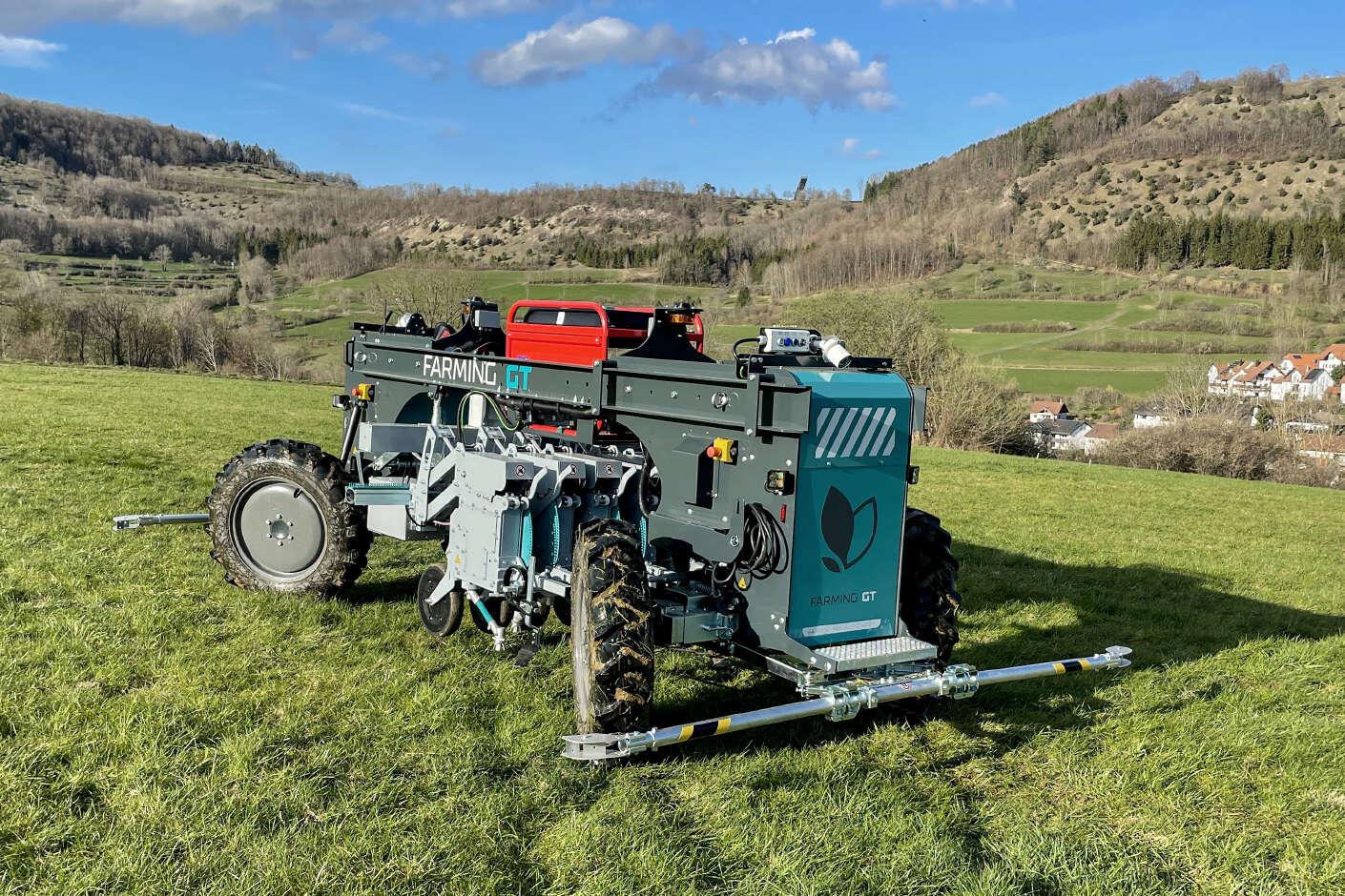 Farming GT steht einsatzbereit auf Grünland in hügeliger Landschaft mit Dorf im Hintergrund