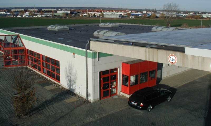 Gewerbehalle mit großem Eingangsbereich, Flachdach und befestigtem Parkplatz, im Hintergrund Industriegebiet.