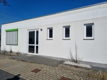 Modernes, eingeschossiges Gebäude mit weißer Putzfassade, drei kleinen Fenstern und Glaseingangstür.