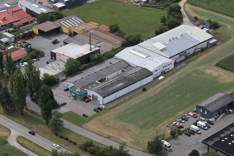 Luftaufnahme eines weitläufigen Gewerbe- und Industrieareals mit mehreren Hallen, Bürogebäuden, Parkflächen und angrenzenden Grünflächen.