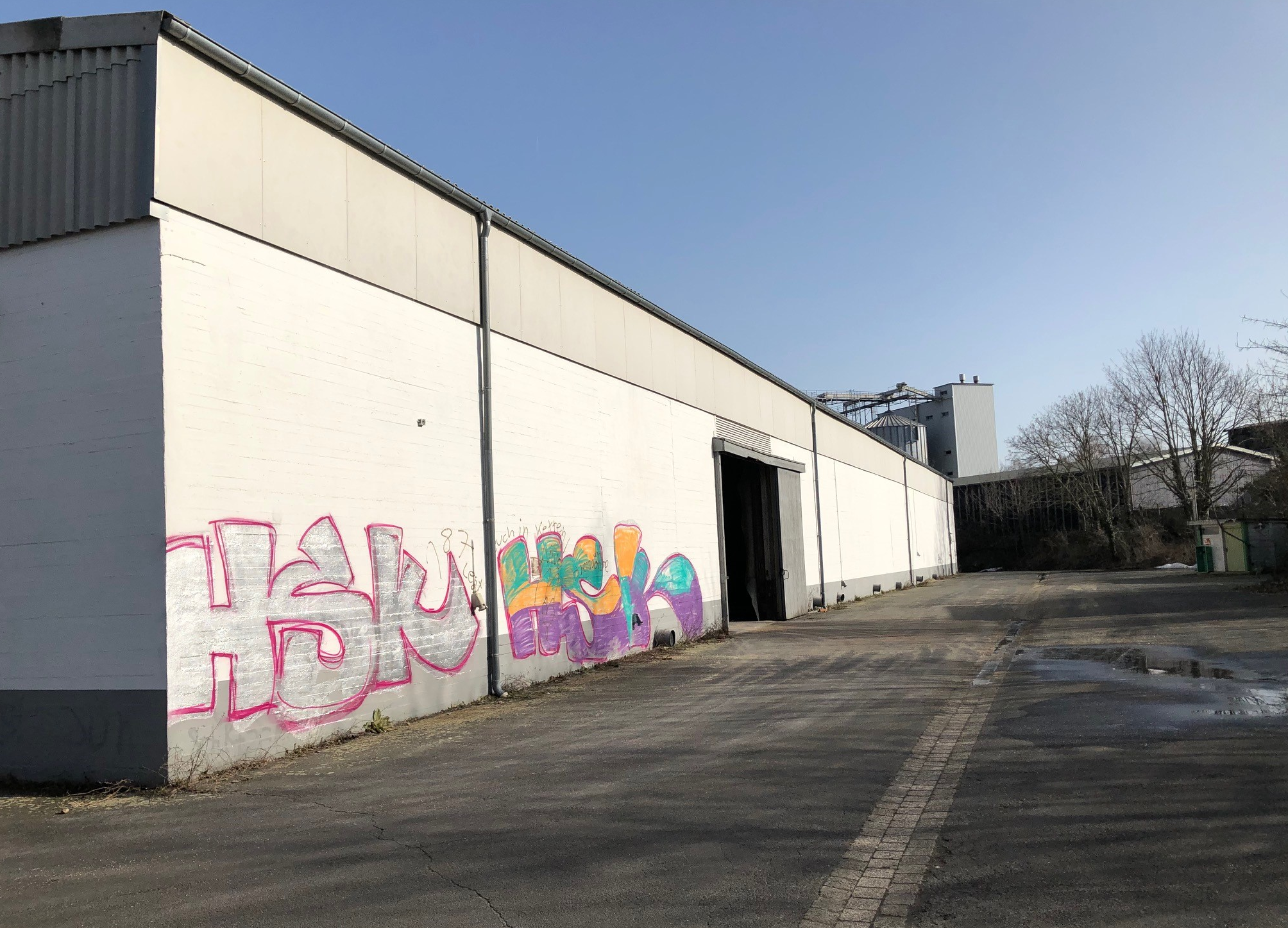 Lange Hallenaußenwand mit geöffnetem Tor und leerem Vorplatz bei Sonnenschein