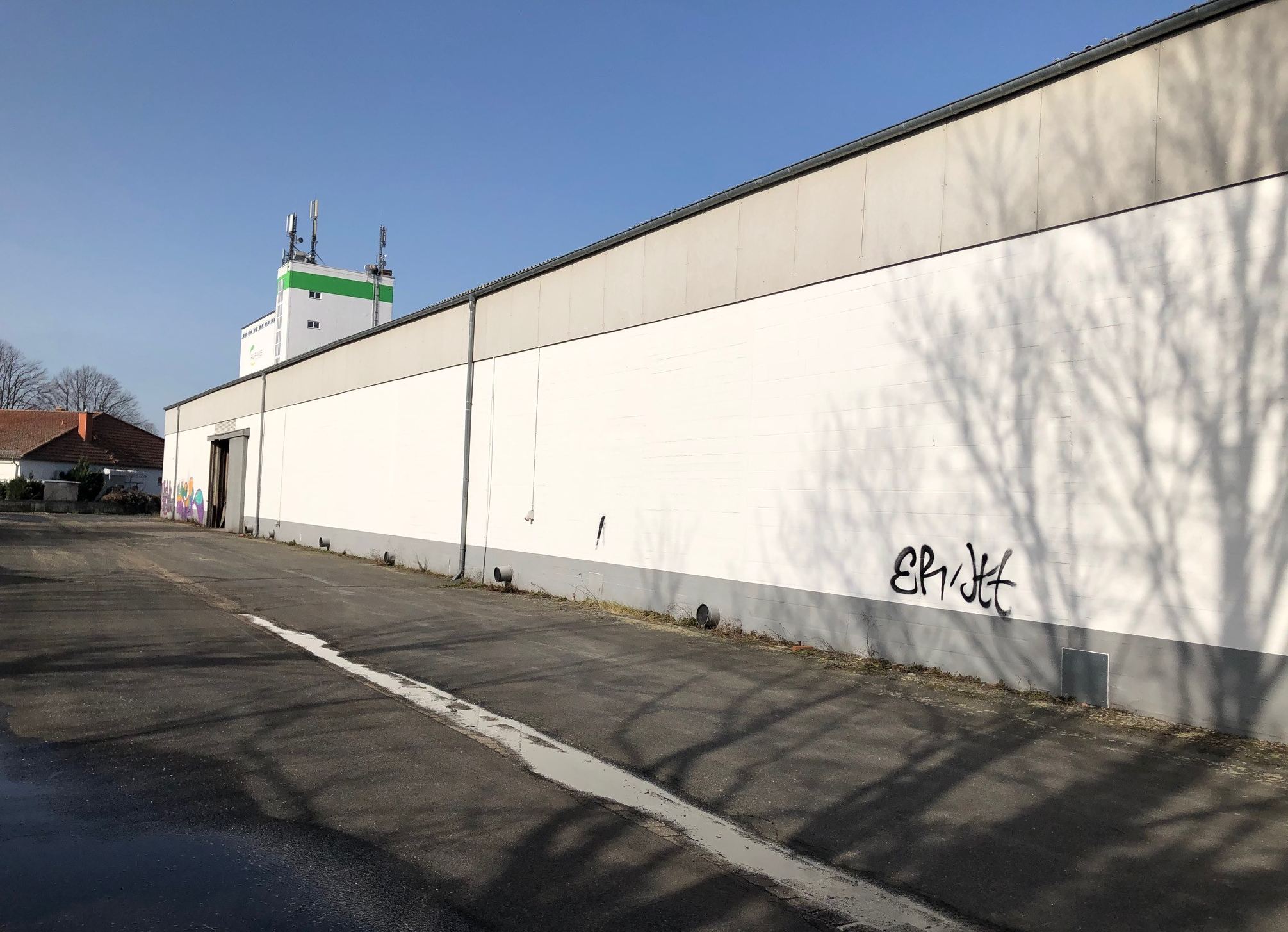 Lange, helle Hallenfassade mit Schatten von Bäumen auf leerem Vorplatz