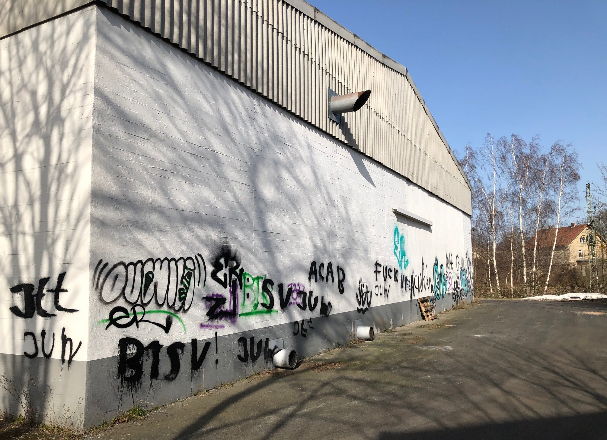 Seitenansicht einer Industriehalle bei Sonnenschein mit angrenzendem Asphaltweg und Bäumen im Hinter