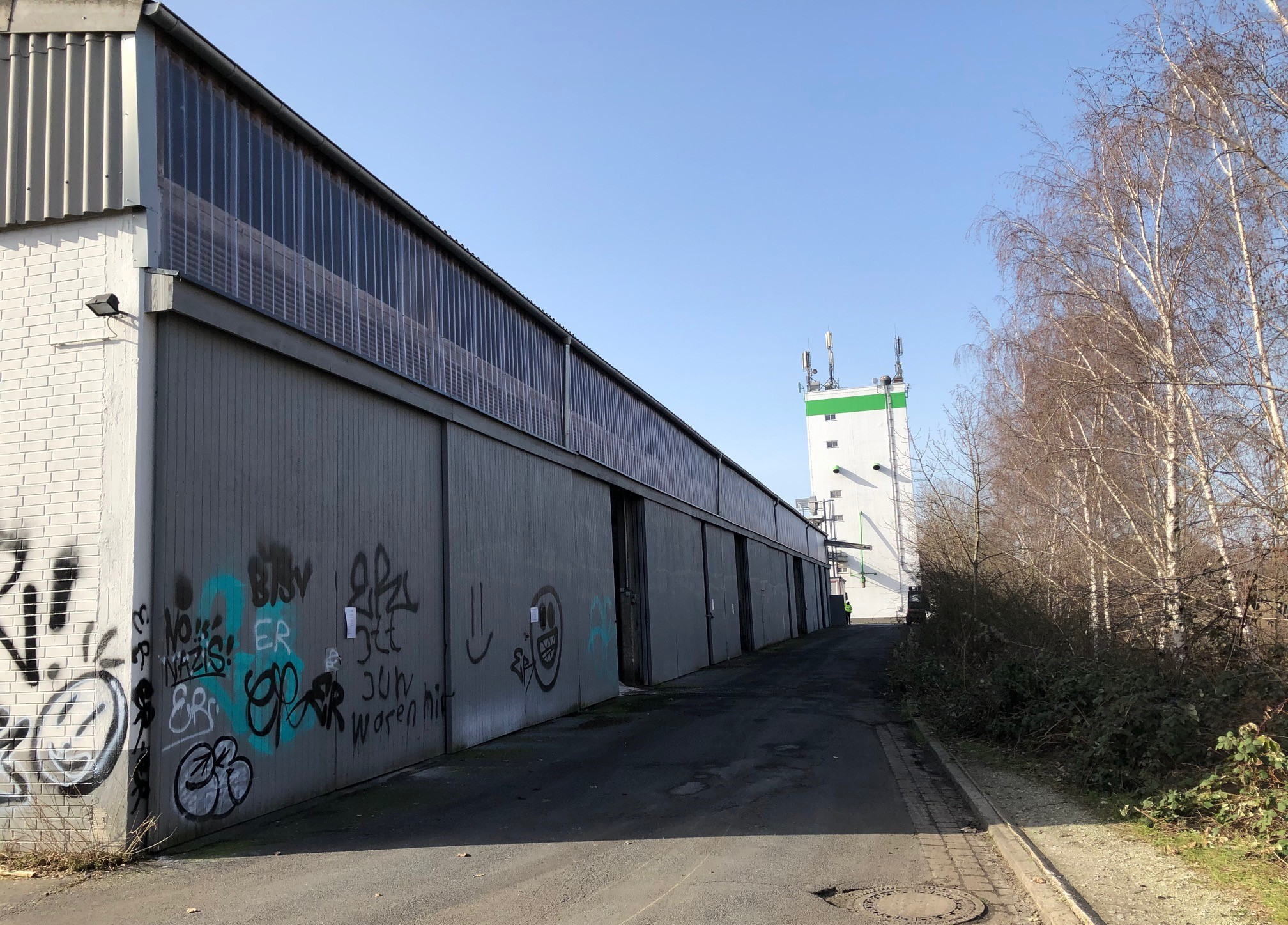 Lange Industriehalle mit geöffneten Toren an einem asphaltierten Weg, rechts Bäume und Sträucher bei