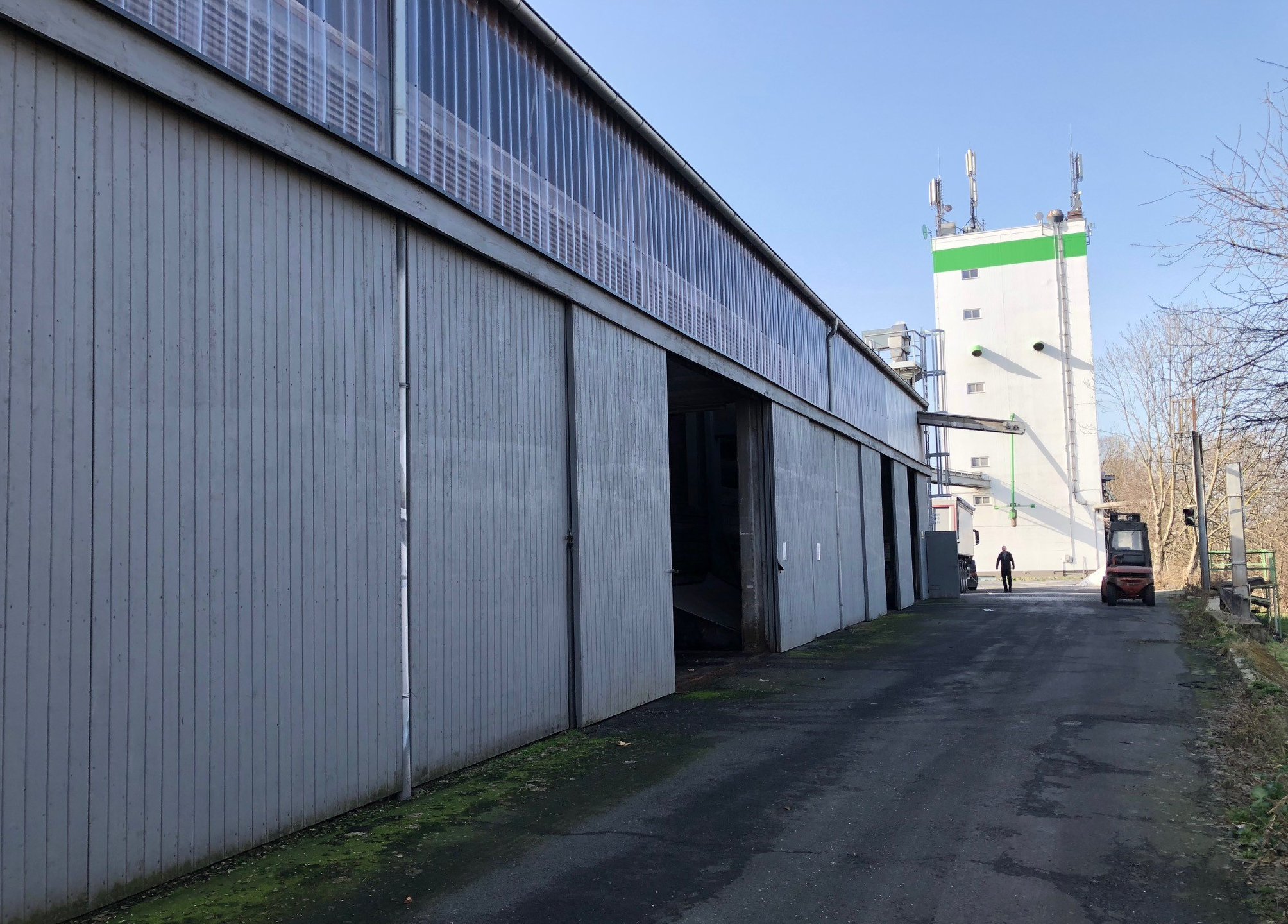 Industriehalle mit geöffnetem Tor an einem asphaltierten Weg, im Hintergrund ein hoher Gebäudeturm u