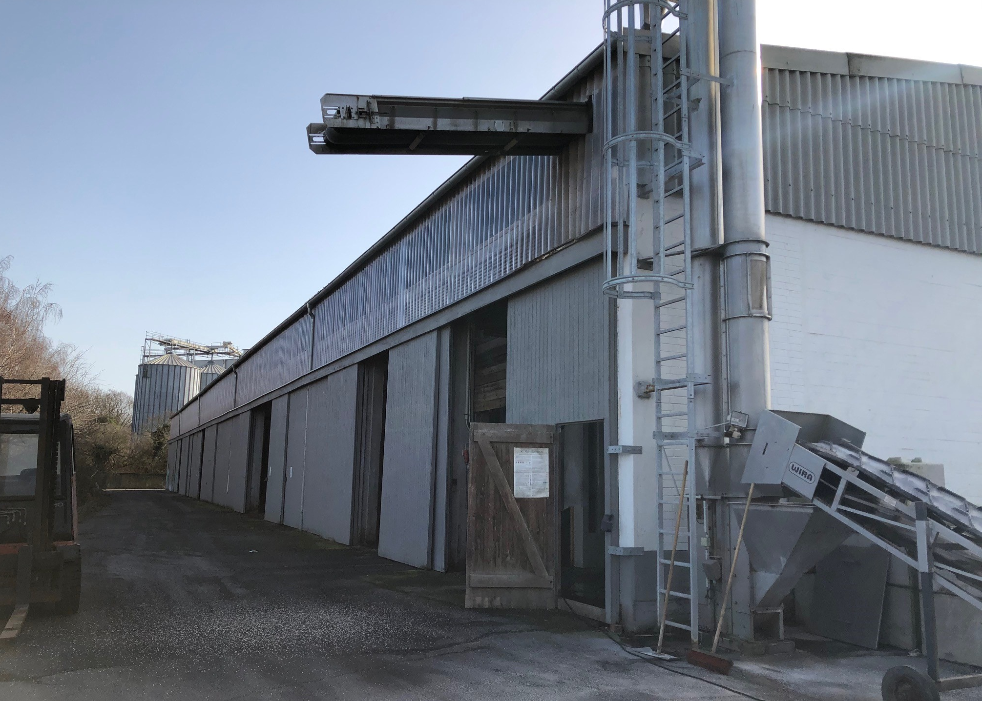 Industriehalle mit mehreren geöffneten Toren, Förderanlage im Vordergrund und Zufahrtsweg entlang de