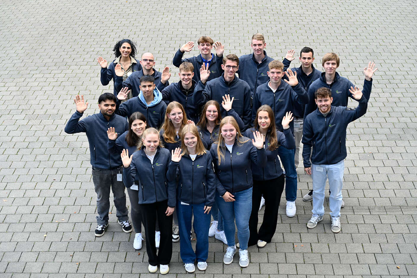 Eine Gruppe von 20 jungen Frauen und Männern stehen auf einem Hof und winken in die Kamera