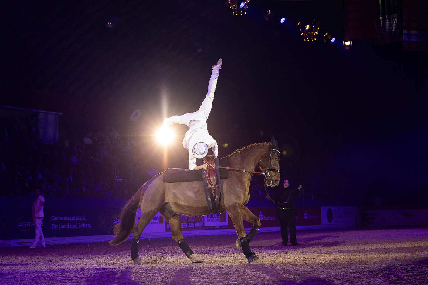 Mann im Kostüm macht einen Handstand auf einem reitenden Pferd im Parcours