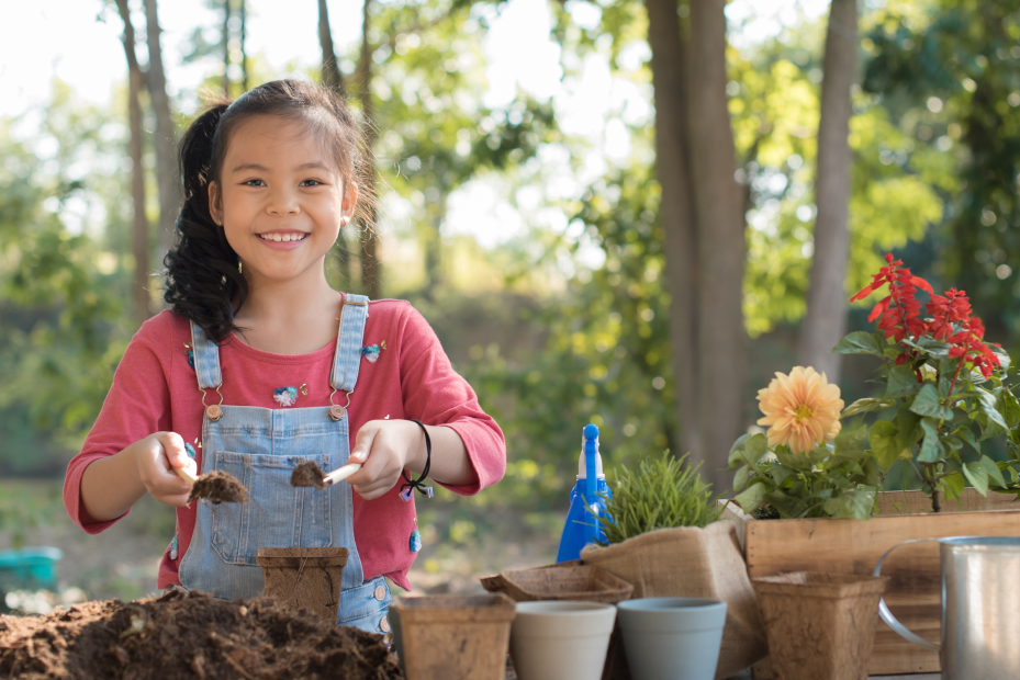 Ein Kind hält zwei kleine Schippen mit Erde. Es ist umgeben von Blumentöpfen, Pflanzen und Erde.
