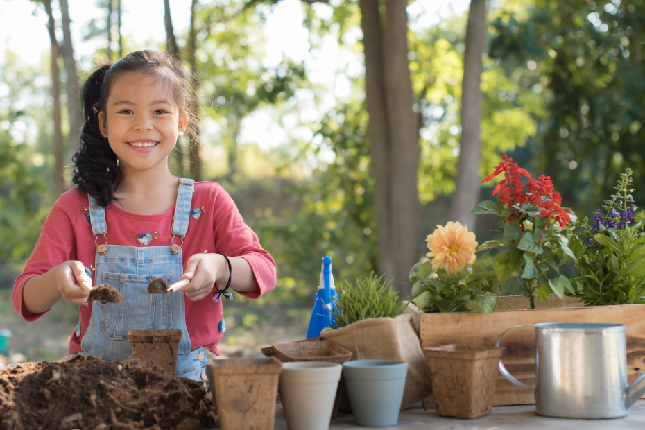 Ein Kind hält zwei kleine Schippen mit Erde. Es ist umgeben von Blumentöpfen, Pflanzen und Erde.