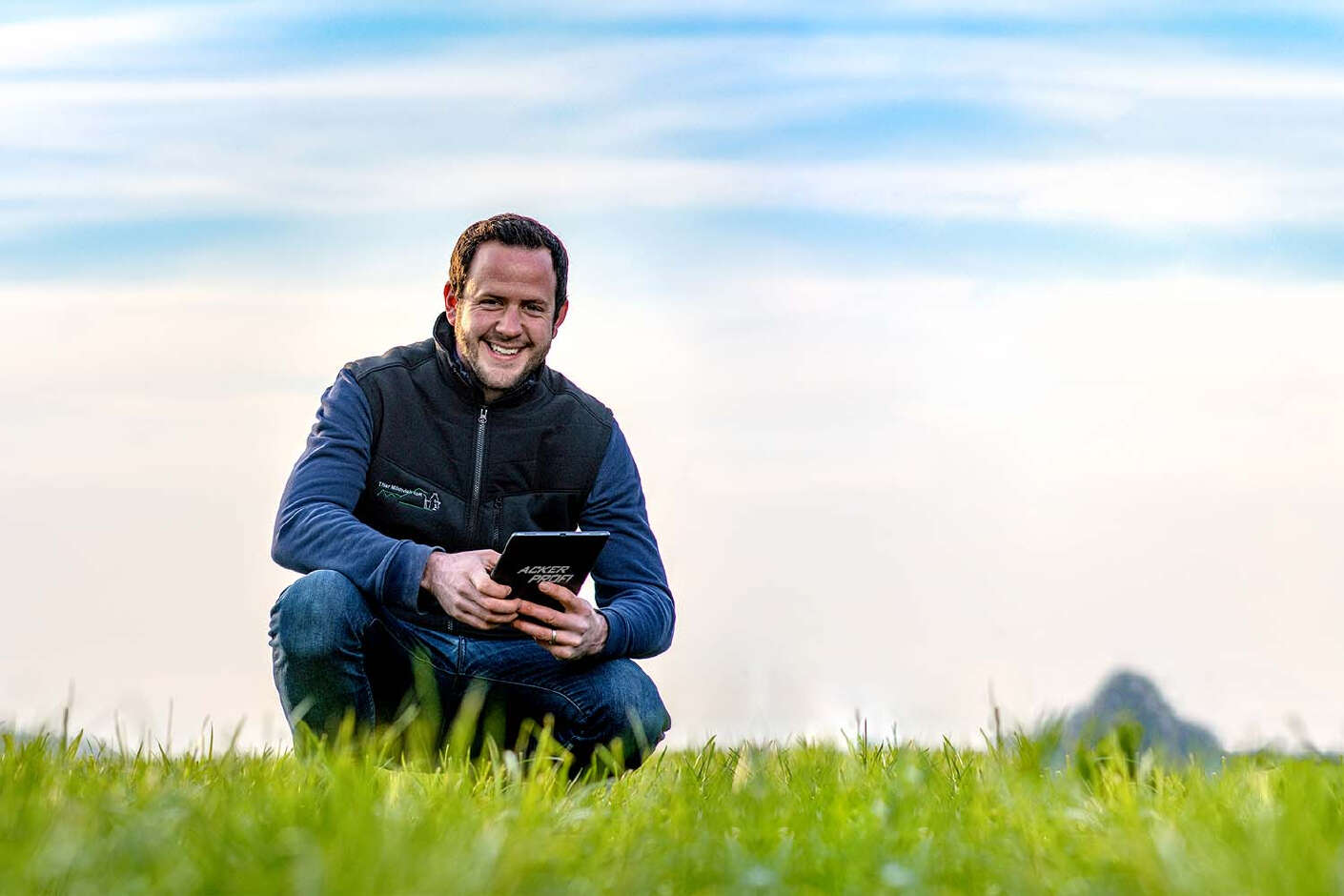 Ein Mann mit einem IPad hockt auf einer Grünlandfläche.