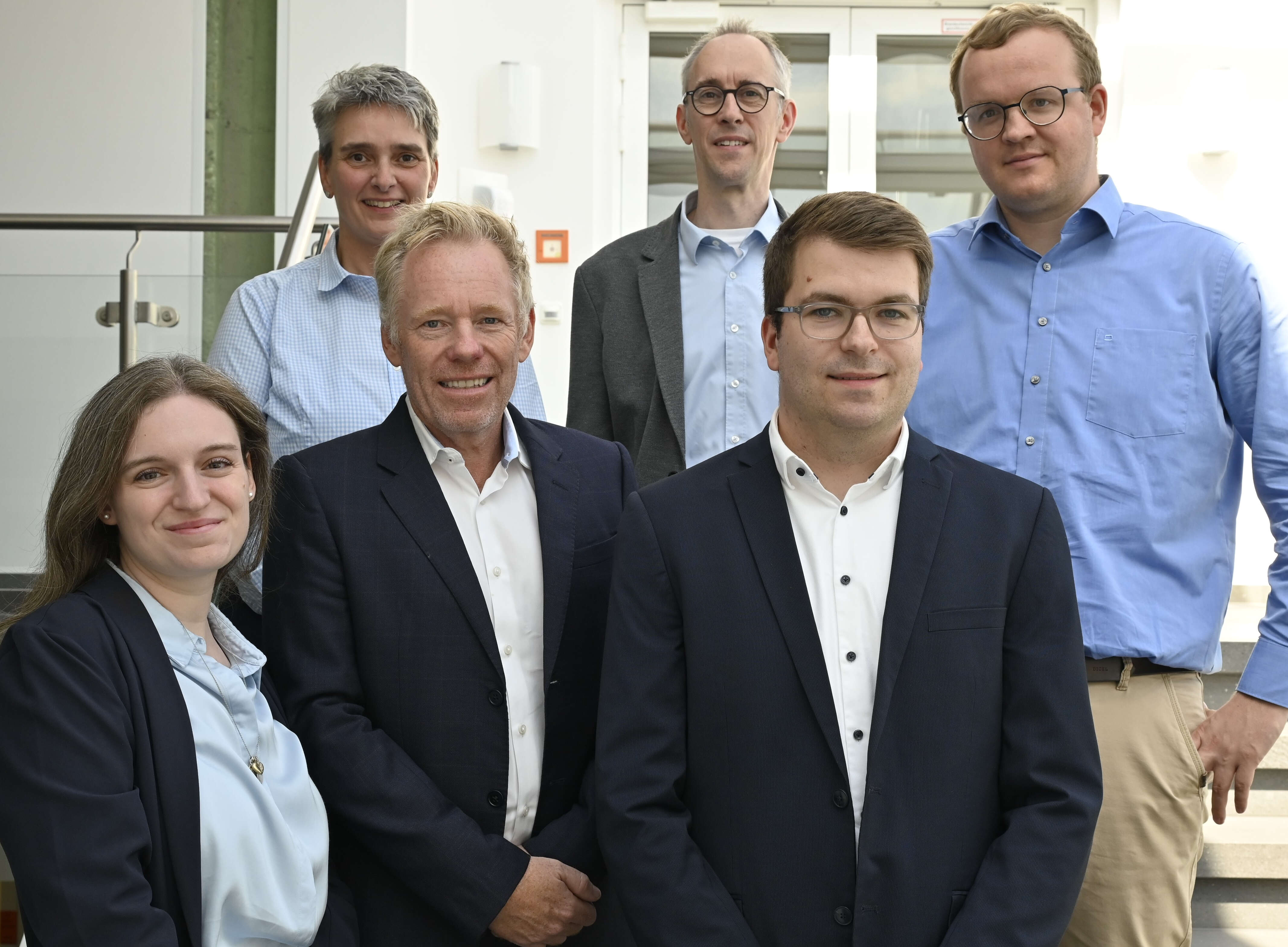 Auf dem Bild stehen sechs Menschen in Businesskleidung vor einem hellen Hintergrund.