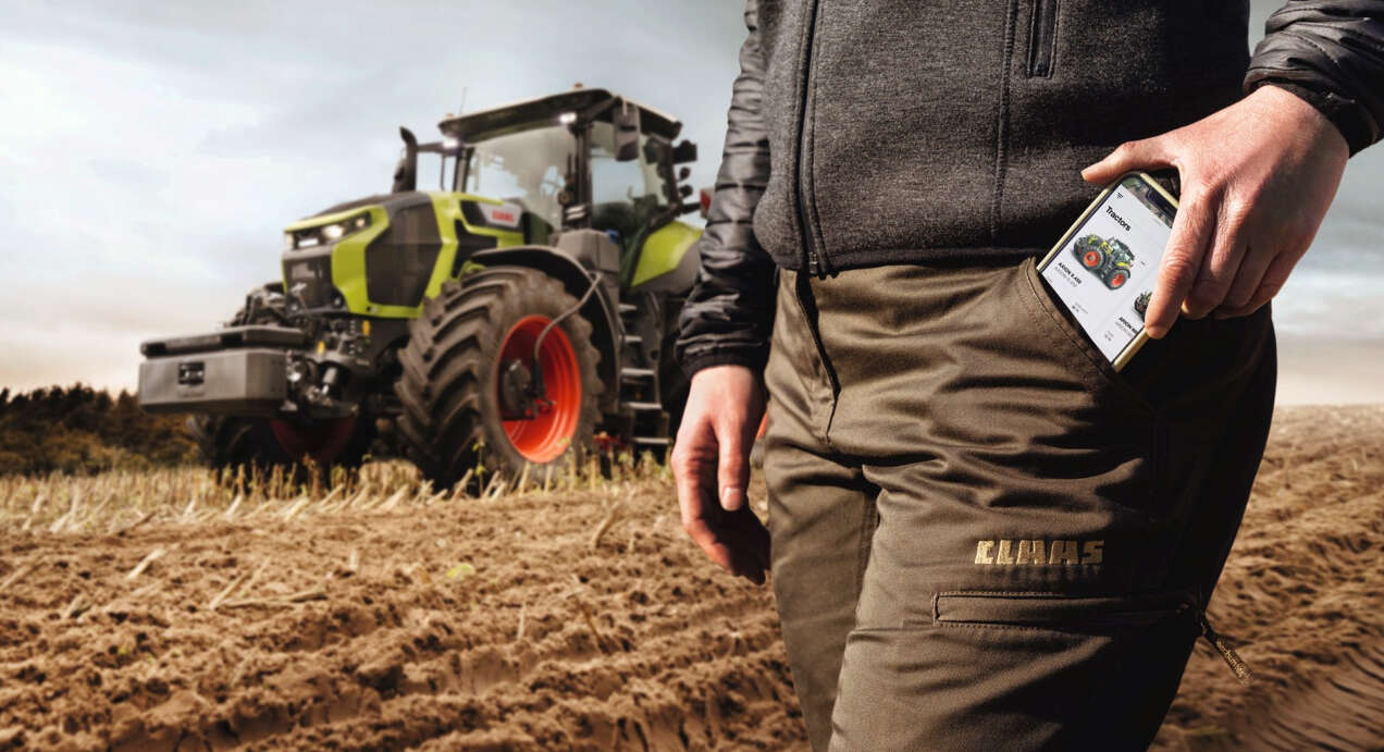Person auf einem Stoppelfeld schiebt ihr Handy in ihre Hosentasche, Claas-Traktor im Hintergrund