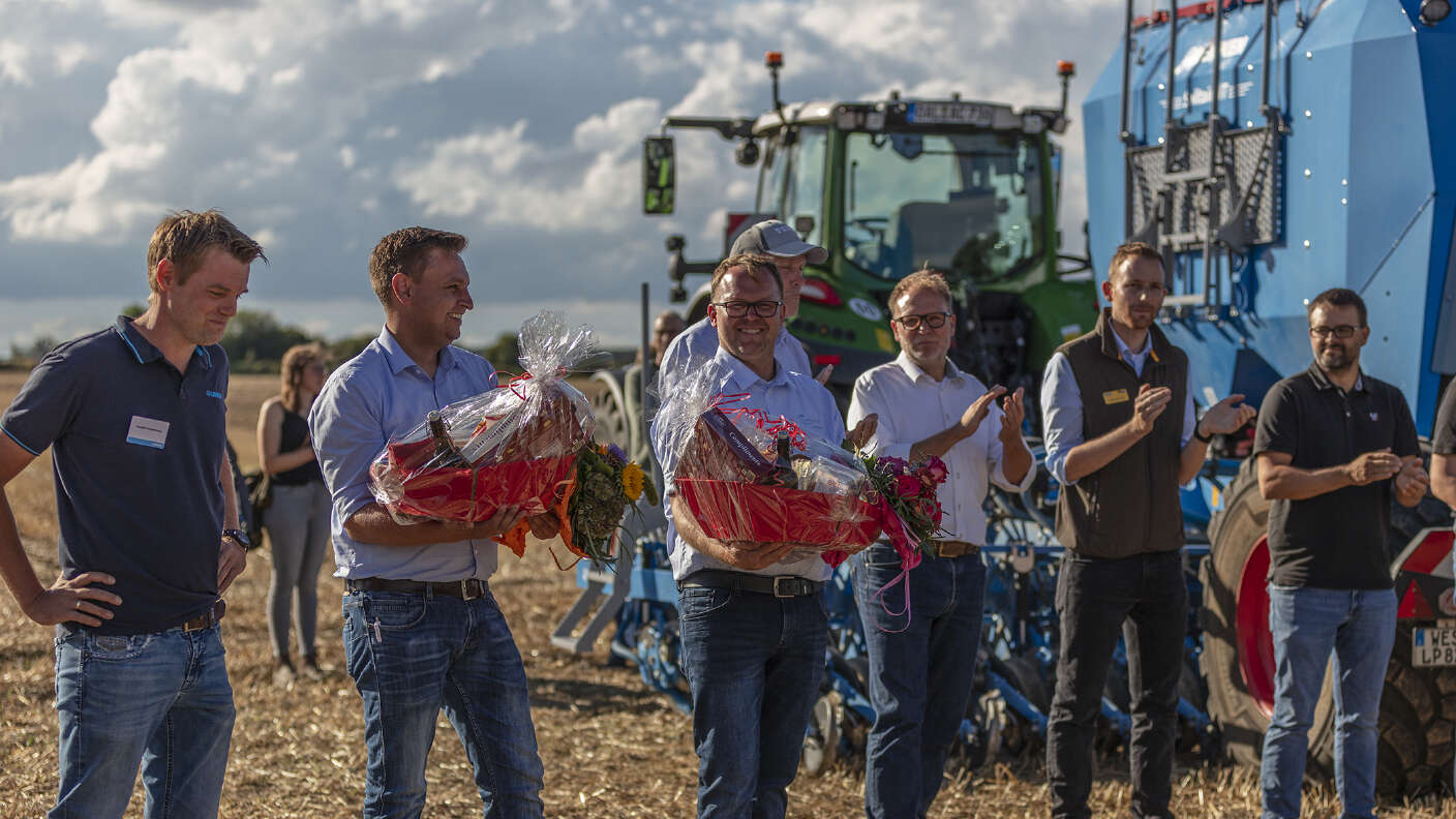 Übergabe von Blumenpräsenten vor Landtechnik auf einer Feldveranstaltung.