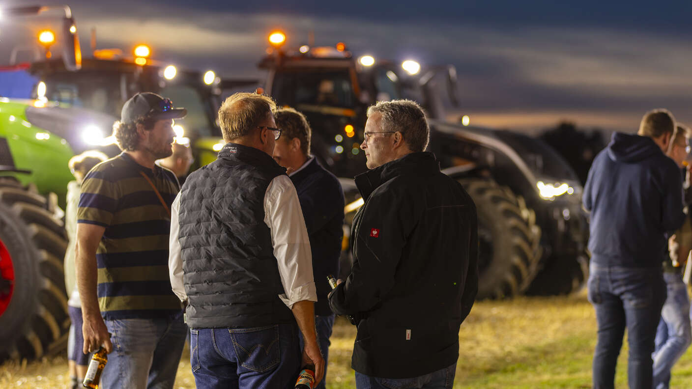 Besucher im Gespräch vor beleuchteten Fendt Traktoren auf einem Stoppelfeld bei Abenddämmerung.
