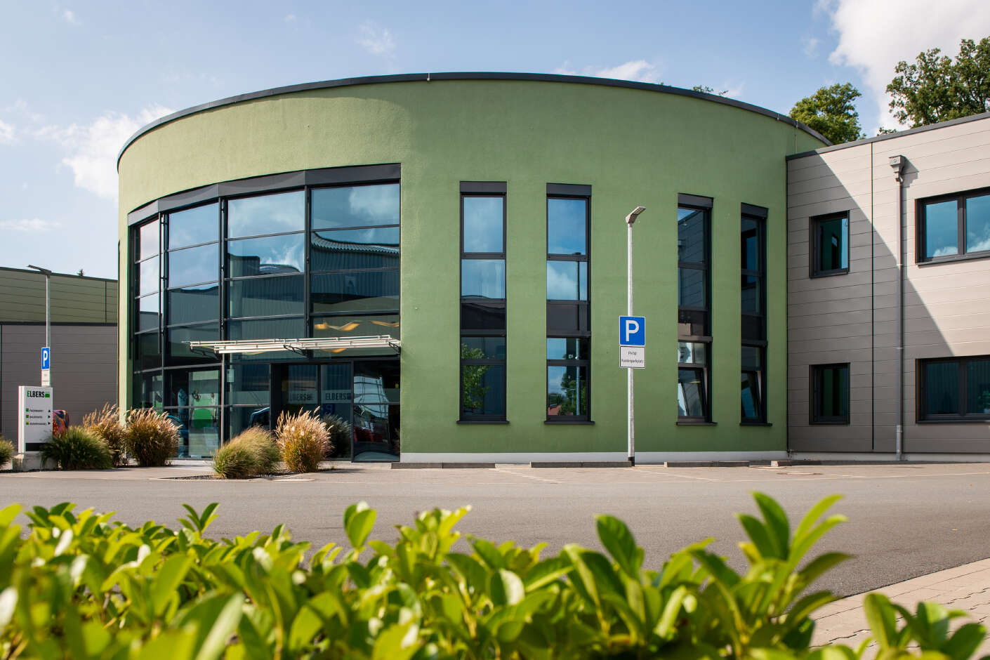 Zweistöckiges, rundes Gebäude mit grüner Putzfassade und dunklen, verspiegelten, großen Fenstern. 